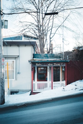 A small storefront with a rustic charm, featuring a red door and white trim. The sign above the entrance reads 'Herbs Garden Thing.' The building is nestled next to a snowy sidewalk on a sloping street, with bare trees in the background.