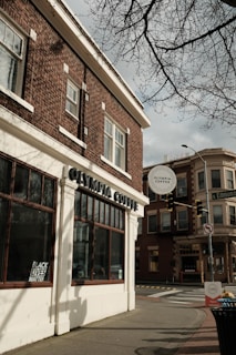 A street corner featuring a brick building with large windows, displaying a sign for Olympia Coffee. A 'Black Lives Matter' poster is visible in one of the windows. Across the street, another building with similar architectural style is present, and traffic lights are part of the urban setting. The sky is overcast, and tree branches are visible at the top.
