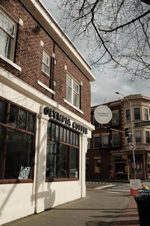 A street corner featuring a brick building with large windows, displaying a sign for Olympia Coffee. A 'Black Lives Matter' poster is visible in one of the windows. Across the street, another building with similar architectural style is present, and traffic lights are part of the urban setting. The sky is overcast, and tree branches are visible at the top.