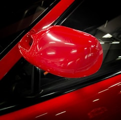 A shiny red side mirror of a car is prominently featured. The background includes a portion of the vehicle's door and window with reflections of overhead lighting.