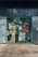 A collection of colorful advertisements and signs covering a metal gate entrance. The signs include promotions for beverages, with various prices and brands displayed. The background shows a concrete sidewalk and a building exterior with bars and a glass window.
