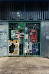 A collection of colorful advertisements and signs covering a metal gate entrance. The signs include promotions for beverages, with various prices and brands displayed. The background shows a concrete sidewalk and a building exterior with bars and a glass window.