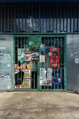 A collection of colorful advertisements and signs covering a metal gate entrance. The signs include promotions for beverages, with various prices and brands displayed. The background shows a concrete sidewalk and a building exterior with bars and a glass window.