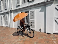 A person riding a bicycle on a brick path alongside a white building with shutters. The cyclist holds an orange umbrella for shade, attached to the bicycle, creating an unusual and eye-catching sight. The umbrella has writing on it, suggesting ownership or instructions.