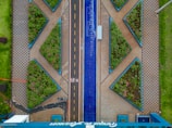 Aerial view of an urban area featuring geometric patterns with tiled walkways and triangular gardens filled with greenery. A central road divides the space, with a blue decorative element running parallel alongside it. A bicycle path marked with symbols is visible, with a parked bicycle present near the bottom.