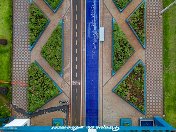 Aerial view of an urban area featuring geometric patterns with tiled walkways and triangular gardens filled with greenery. A central road divides the space, with a blue decorative element running parallel alongside it. A bicycle path marked with symbols is visible, with a parked bicycle present near the bottom.