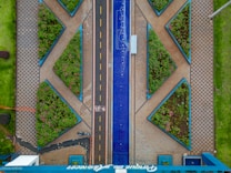 Aerial view of an urban area featuring geometric patterns with tiled walkways and triangular gardens filled with greenery. A central road divides the space, with a blue decorative element running parallel alongside it. A bicycle path marked with symbols is visible, with a parked bicycle present near the bottom.