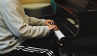 A person in a light-colored jacket and striped athletic pants is playing a grand piano, focusing on the keys. The setting appears to be indoors with subdued lighting, creating a warm and intimate atmosphere.