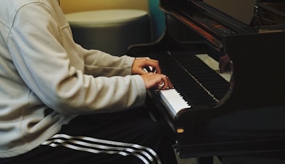 A person in a light-colored jacket and striped athletic pants is playing a grand piano, focusing on the keys. The setting appears to be indoors with subdued lighting, creating a warm and intimate atmosphere.