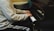 A person in a light-colored jacket and striped athletic pants is playing a grand piano, focusing on the keys. The setting appears to be indoors with subdued lighting, creating a warm and intimate atmosphere.