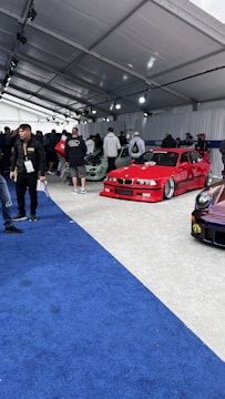 A car exhibition with several modified cars displayed under a large white tent. The prominently featured car is a bright red BMW with sporty modifications. Several people are gathered around, some inspecting the cars and others engaged in conversation. The ground is covered with blue and gray carpeting, and bright overhead lights illuminate the scene.
