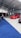 A car exhibition with several modified cars displayed under a large white tent. The prominently featured car is a bright red BMW with sporty modifications. Several people are gathered around, some inspecting the cars and others engaged in conversation. The ground is covered with blue and gray carpeting, and bright overhead lights illuminate the scene.