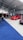 A car exhibition with several modified cars displayed under a large white tent. The prominently featured car is a bright red BMW with sporty modifications. Several people are gathered around, some inspecting the cars and others engaged in conversation. The ground is covered with blue and gray carpeting, and bright overhead lights illuminate the scene.