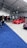 A car exhibition with several modified cars displayed under a large white tent. The prominently featured car is a bright red BMW with sporty modifications. Several people are gathered around, some inspecting the cars and others engaged in conversation. The ground is covered with blue and gray carpeting, and bright overhead lights illuminate the scene.
