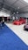 A car exhibition with several modified cars displayed under a large white tent. The prominently featured car is a bright red BMW with sporty modifications. Several people are gathered around, some inspecting the cars and others engaged in conversation. The ground is covered with blue and gray carpeting, and bright overhead lights illuminate the scene.
