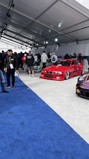 A car exhibition with several modified cars displayed under a large white tent. The prominently featured car is a bright red BMW with sporty modifications. Several people are gathered around, some inspecting the cars and others engaged in conversation. The ground is covered with blue and gray carpeting, and bright overhead lights illuminate the scene.