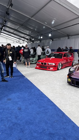 A car exhibition with several modified cars displayed under a large white tent. The prominently featured car is a bright red BMW with sporty modifications. Several people are gathered around, some inspecting the cars and others engaged in conversation. The ground is covered with blue and gray carpeting, and bright overhead lights illuminate the scene.