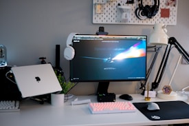 A neatly arranged computer workstation featuring a monitor with a vibrant, abstract screensaver. A laptop with a logo is placed on the side, accompanied by a mechanical keyboard with backlighting and a basic mouse on a large mousepad. Over-ear headphones rest on top of the monitor, and a desk lamp provides lighting from the right. The background features a pegboard with various accessories and headphones hanging on it.