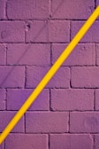 A textured brick wall painted in deep purple is intersected diagonally by a smooth, bright yellow pipe.