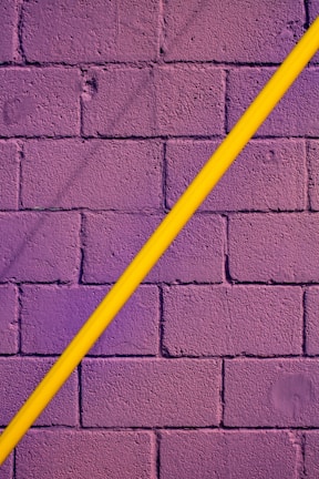 A textured brick wall painted in deep purple is intersected diagonally by a smooth, bright yellow pipe.