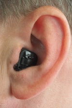 A close-up view of a human ear with an in-ear hearing aid device in place. The hearing aid is black, fitting snugly within the contours of the ear.