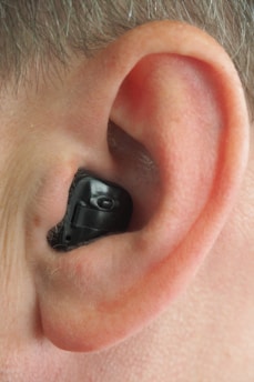 A close-up view of a human ear with an in-ear hearing aid device in place. The hearing aid is black, fitting snugly within the contours of the ear.