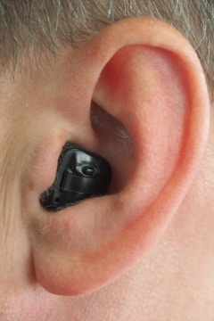 A close-up view of a human ear with an in-ear hearing aid device in place. The hearing aid is black, fitting snugly within the contours of the ear.