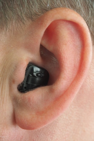 A close-up view of a human ear with an in-ear hearing aid device in place. The hearing aid is black, fitting snugly within the contours of the ear.
