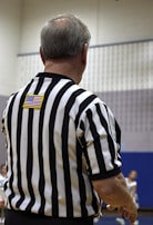 A person in a black and white striped referee shirt is standing with their back to the camera. The American flag patch is visible on the upper back. The background is a gymnasium with blurred figures and a basketball game in progress.