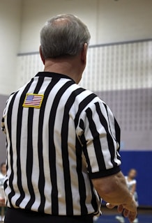 A person in a black and white striped referee shirt is standing with their back to the camera. The American flag patch is visible on the upper back. The background is a gymnasium with blurred figures and a basketball game in progress.