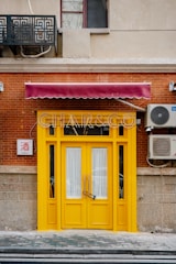 A bright yellow door with glass panels set in a red brick wall, adorned by a burgundy awning. There is an air conditioning unit mounted on the wall to the right and a decorative metal grate above. A small sign with Chinese characters is visible on the left.