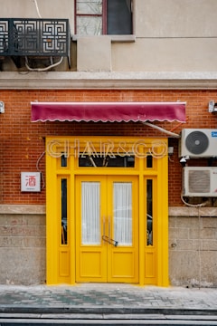 A bright yellow door with glass panels set in a red brick wall, adorned by a burgundy awning. There is an air conditioning unit mounted on the wall to the right and a decorative metal grate above. A small sign with Chinese characters is visible on the left.