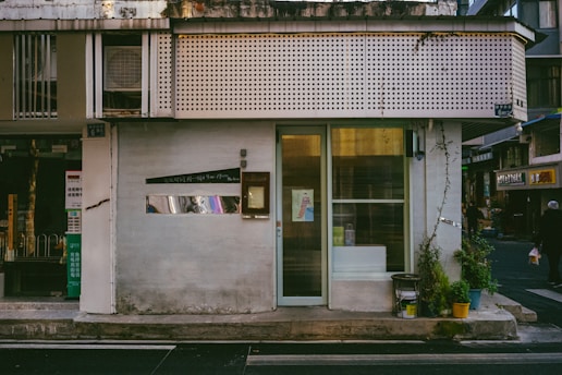A small, storefront with a minimalist design. The exterior features a light-colored facade with a vertical glass window and door. Potted plants are arranged neatly by the entrance. An air conditioning unit is visible on the upper section of the building. There are signs with Asian script, indicating a possible location in an Asian country. The adjacent street has a few small shops and a passerby.