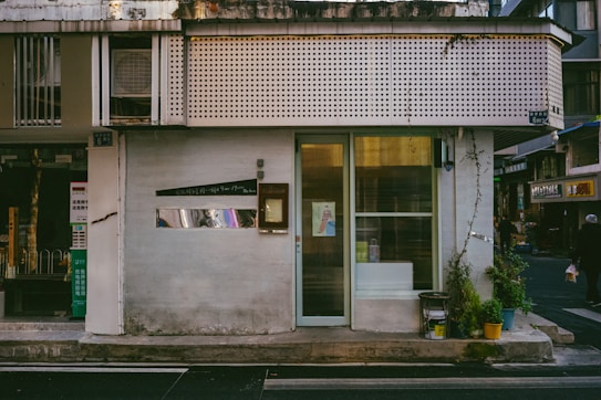 A small, storefront with a minimalist design. The exterior features a light-colored facade with a vertical glass window and door. Potted plants are arranged neatly by the entrance. An air conditioning unit is visible on the upper section of the building. There are signs with Asian script, indicating a possible location in an Asian country. The adjacent street has a few small shops and a passerby.