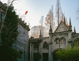 A Gothic-style building with arched windows and pointed spires is surrounded by tall, bare trees. A distant modern apartment building is visible in the background. A red flag is caught in the branches of one of the trees.