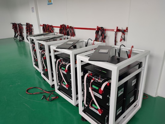 Several battery storage systems are organized in a room with a green floor. Each system is housed within a white metal frame, featuring black components and various wiring connections. Coiled cables hang on the wall, and some cables are on the floor in front of the systems.