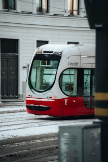 A red and white tram with the name Liepāja on it is seen on a snowy street. The tram is on tracks with snow covering parts of it, and buildings with windows are visible in the background.