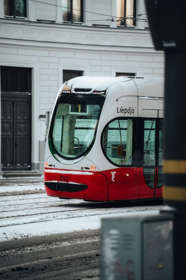 A red and white tram with the name Liepāja on it is seen on a snowy street. The tram is on tracks with snow covering parts of it, and buildings with windows are visible in the background.