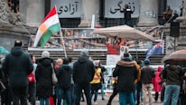A public protest in front of a building with people gathered around a speaker holding a sign that reads 'Stop Chem Attacks on Iranian Girls.' Banners and flags, including a Kurdish flag, are displayed. The scene includes several people dressed in warm clothing due to cold weather, standing and listening.