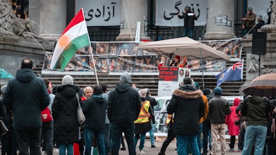 A public protest in front of a building with people gathered around a speaker holding a sign that reads 'Stop Chem Attacks on Iranian Girls.' Banners and flags, including a Kurdish flag, are displayed. The scene includes several people dressed in warm clothing due to cold weather, standing and listening.