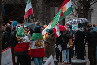 A group of people gathered outdoors, some holding flags and others under umbrellas. Two individuals have flag capes with a lion and sun emblem. The atmosphere appears to be a protest or demonstration.