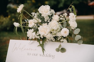 A beautifully arranged bouquet of white flowers, including roses and daisies, is placed on top of a white rectangular sign that reads 'Welcome to our wedding.' The background is a blurred outdoor setting with green foliage.