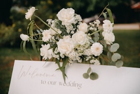 A beautifully arranged bouquet of white flowers, including roses and daisies, is placed on top of a white rectangular sign that reads 'Welcome to our wedding.' The background is a blurred outdoor setting with green foliage.