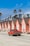A row of charming, identical peach-colored beach houses lines a sandy street, each with steeply pitched roofs. A classic red pickup truck drives past them. The background is dotted with tall palm trees under a bright blue sky.