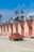 A row of charming, identical peach-colored beach houses lines a sandy street, each with steeply pitched roofs. A classic red pickup truck drives past them. The background is dotted with tall palm trees under a bright blue sky.