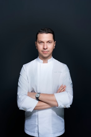 A person stands confidently with arms crossed against a plain dark background, wearing a white chef's jacket. The attire suggests a professional culinary context, and the individual appears focused and composed.
