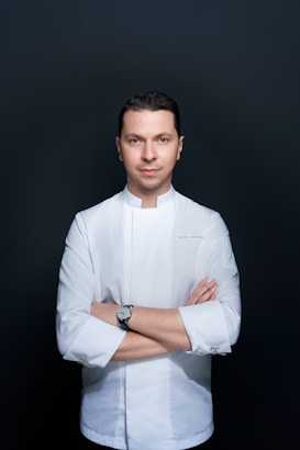 A person stands confidently with arms crossed against a plain dark background, wearing a white chef's jacket. The attire suggests a professional culinary context, and the individual appears focused and composed.