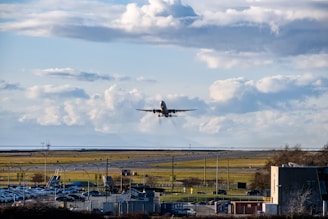 An airplane is taking off from a runway against a backdrop of a cloudy blue sky. The scene includes a wide view of the airport with visible structures, parked vehicles, and grassy areas.