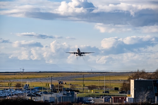 An airplane is taking off from a runway against a backdrop of a cloudy blue sky. The scene includes a wide view of the airport with visible structures, parked vehicles, and grassy areas.