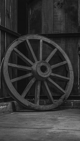 Rustic wooden wagon wheel leaning against a sunlit barn exterior.